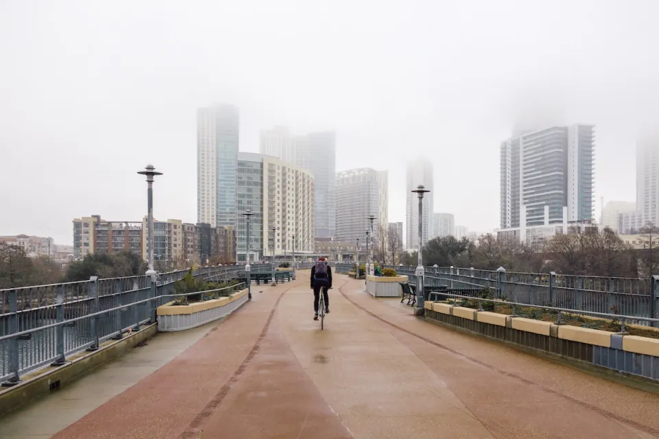 One cyclist rides on pedestrian bridge with foggy Austin, Texas skyline in background.